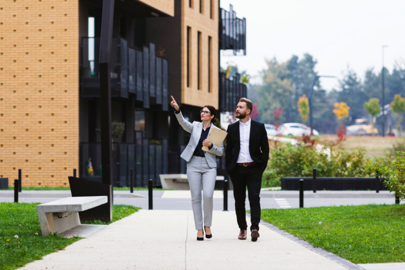 A female real estate agent in a gray blazer and glasses walks alongside a male client in a dark suit along a sidewalk, gesturing toward a modern multi-story residential