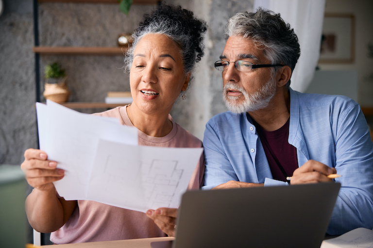 Senior couple analyzing blueprints and using a laptop at home, discussing plans and making decisions together, fostering communication and connection in their partnership